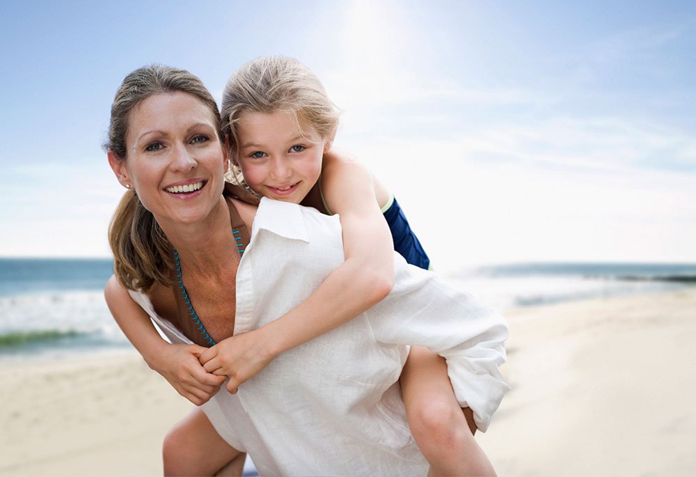 STE mother giving daughter piggyback ride on beach